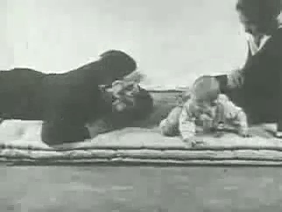 A grainy frame of silent film: a man on all fours wearing a bearded mask leans toward a baby who sits on a white mattress, while a woman reaches in from the right