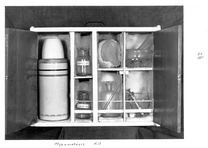 A small wooden case opened to show a thermos flask, labelled bottles, a glass dish and a syringe packed in compartments, with the words Myxomatosis Kit written beneath