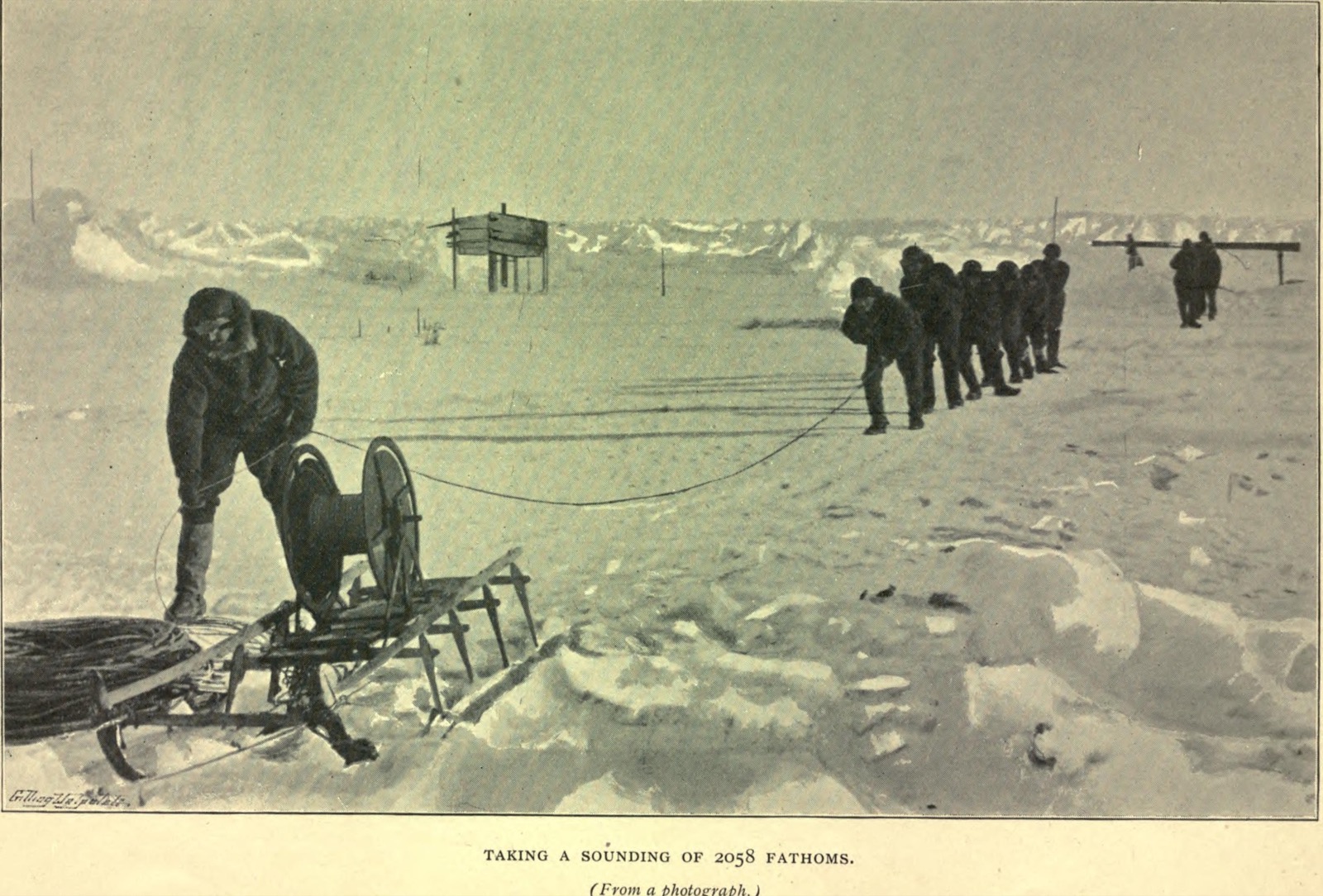 Men on the ice hauling in a line from a hole, one bending over a large reel on a sledge in the foreground, with the ship's masts and a hut in the distance