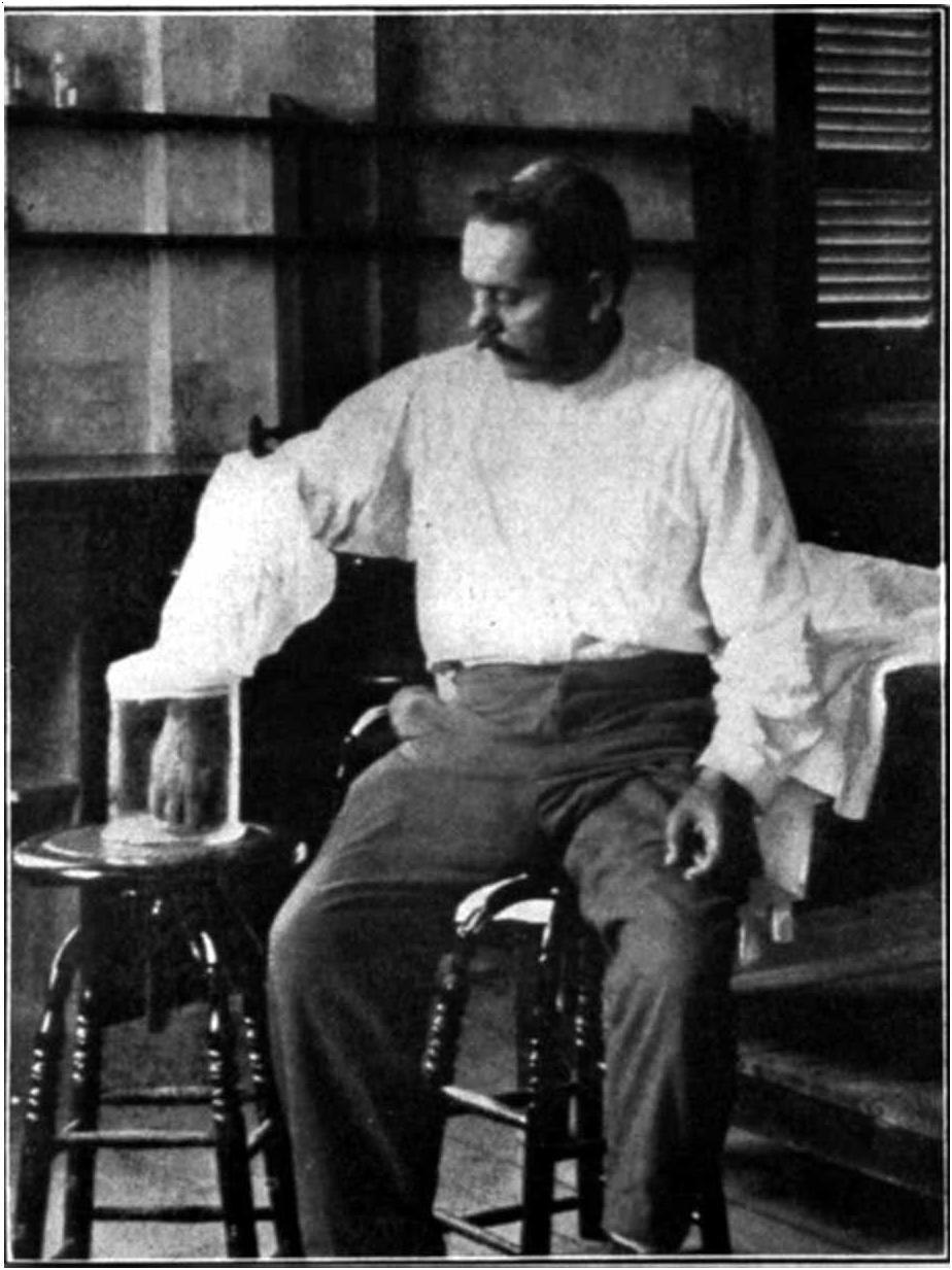 A man in a white shirt sits on a stool with his bare forearm resting on another stool, a glass test tube held upside down against the skin of his arm
