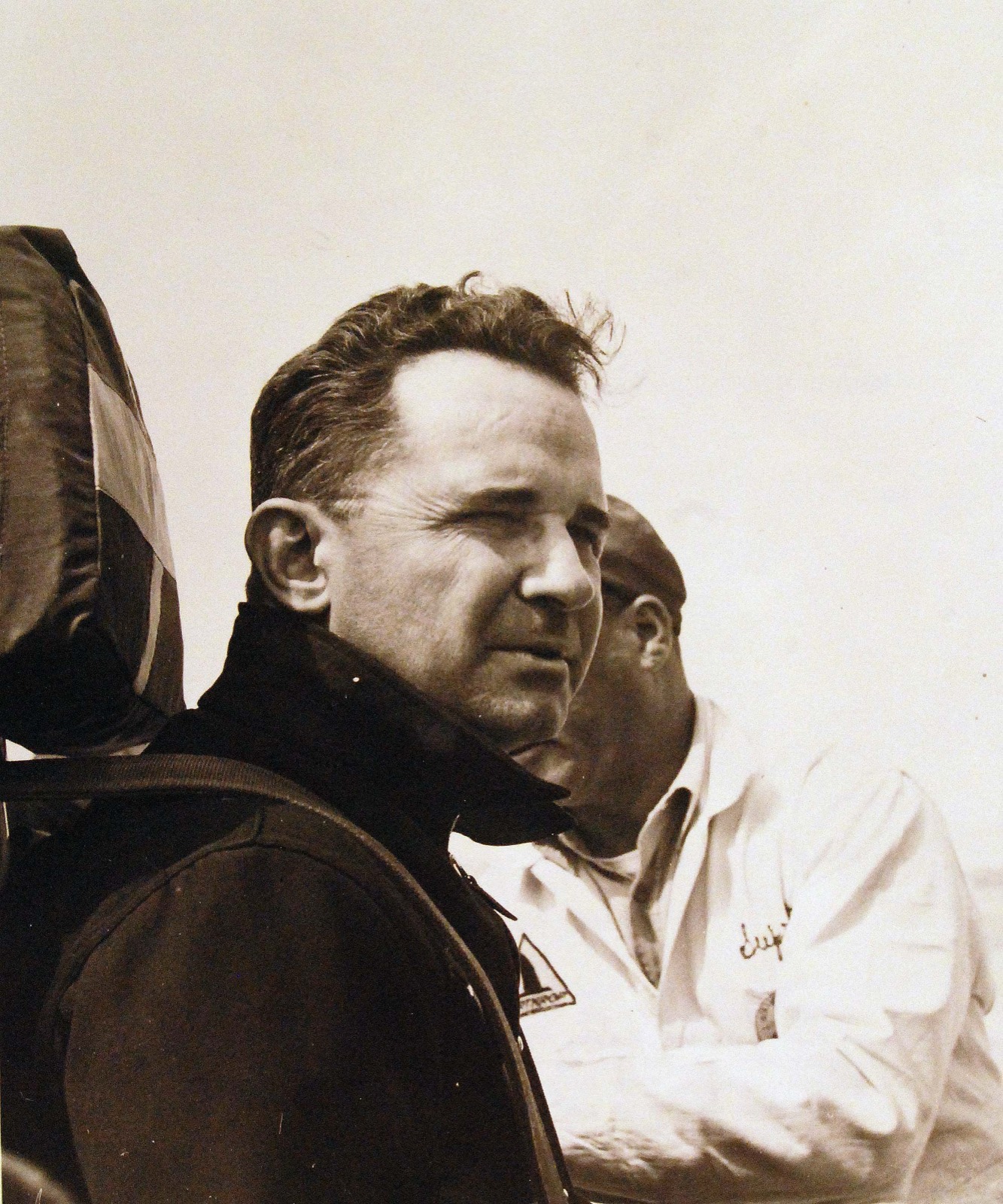 A black and white photograph of a man in a dark flying jacket seen in profile, strapped against the padded headrest of a seat, with a man in white overalls behind him under an empty sky