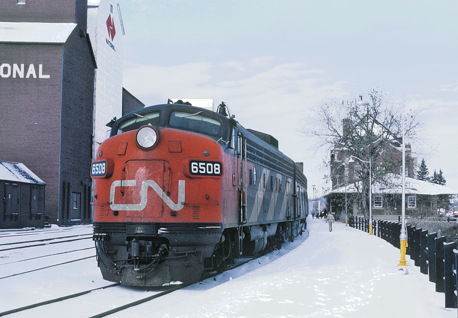 A red and black Canadian National diesel locomotive at a snowy station platform in a small prairie town, grain elevators behind it