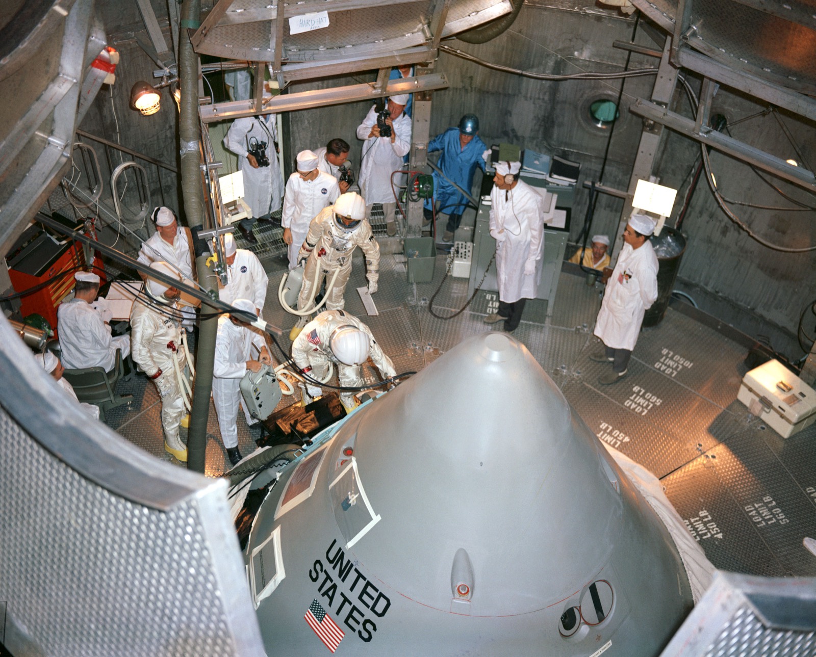 Technicians in white coats and caps stand around the conical top of a spacecraft in a steel-walled altitude chamber while a suited astronaut climbs in through the hatch and another waits behind him