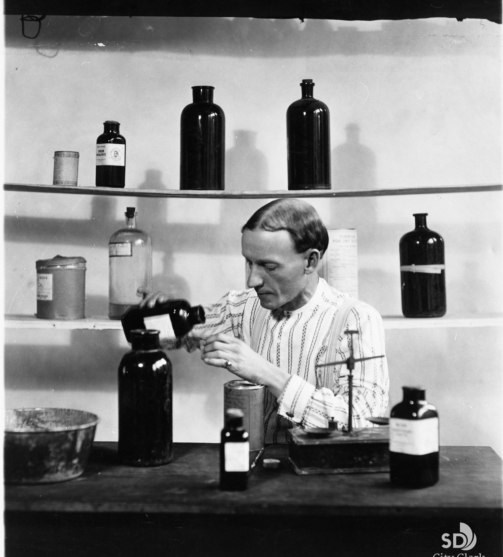 Charles Hatfield in shirtsleeves at a bench, pouring from a dark bottle into a jar, with shelves of bottles and a balance behind him
