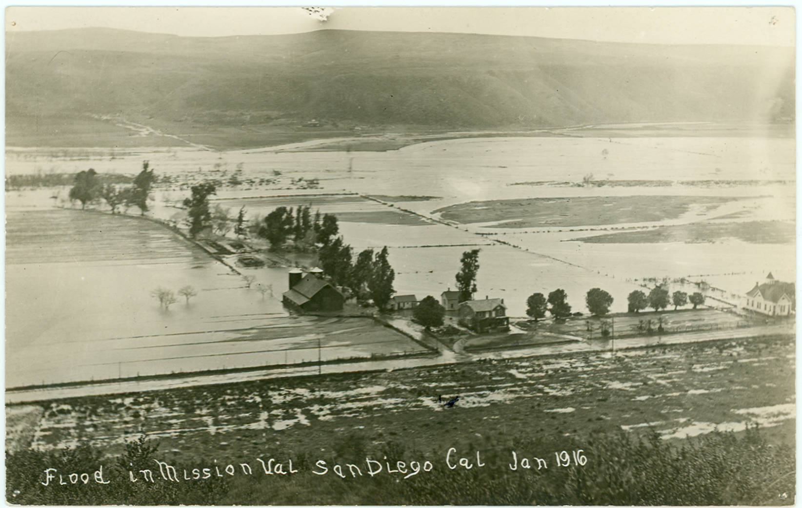 A valley floor under sheets of floodwater with a farmhouse, barn and rows of trees standing in it, seen from a hillside, with the caption written on the negative