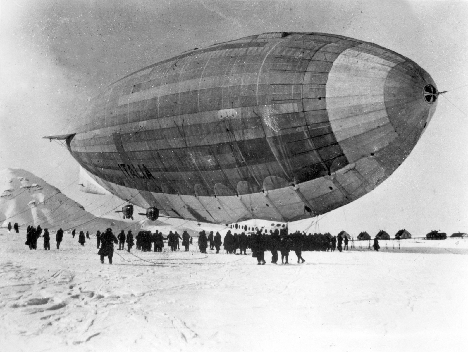 A large grey airship with ITALIA on its flank hangs low over a snowfield while a crowd of men in dark coats hold its ropes, with huts and a mountain behind