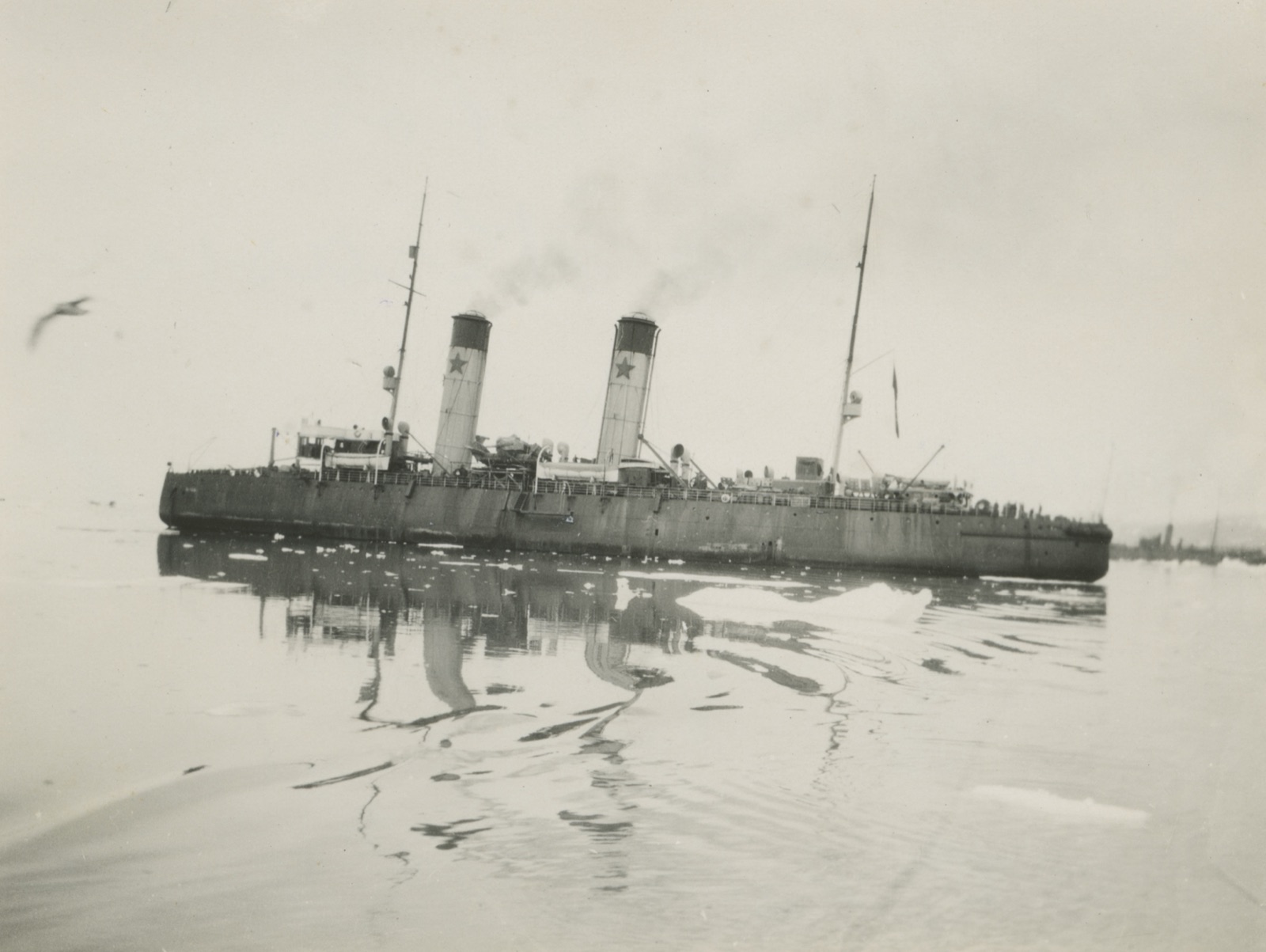 A long dark ice-breaker with two tall funnels, each painted with a star, lies in flat water with small floes around her and a gull in the air
