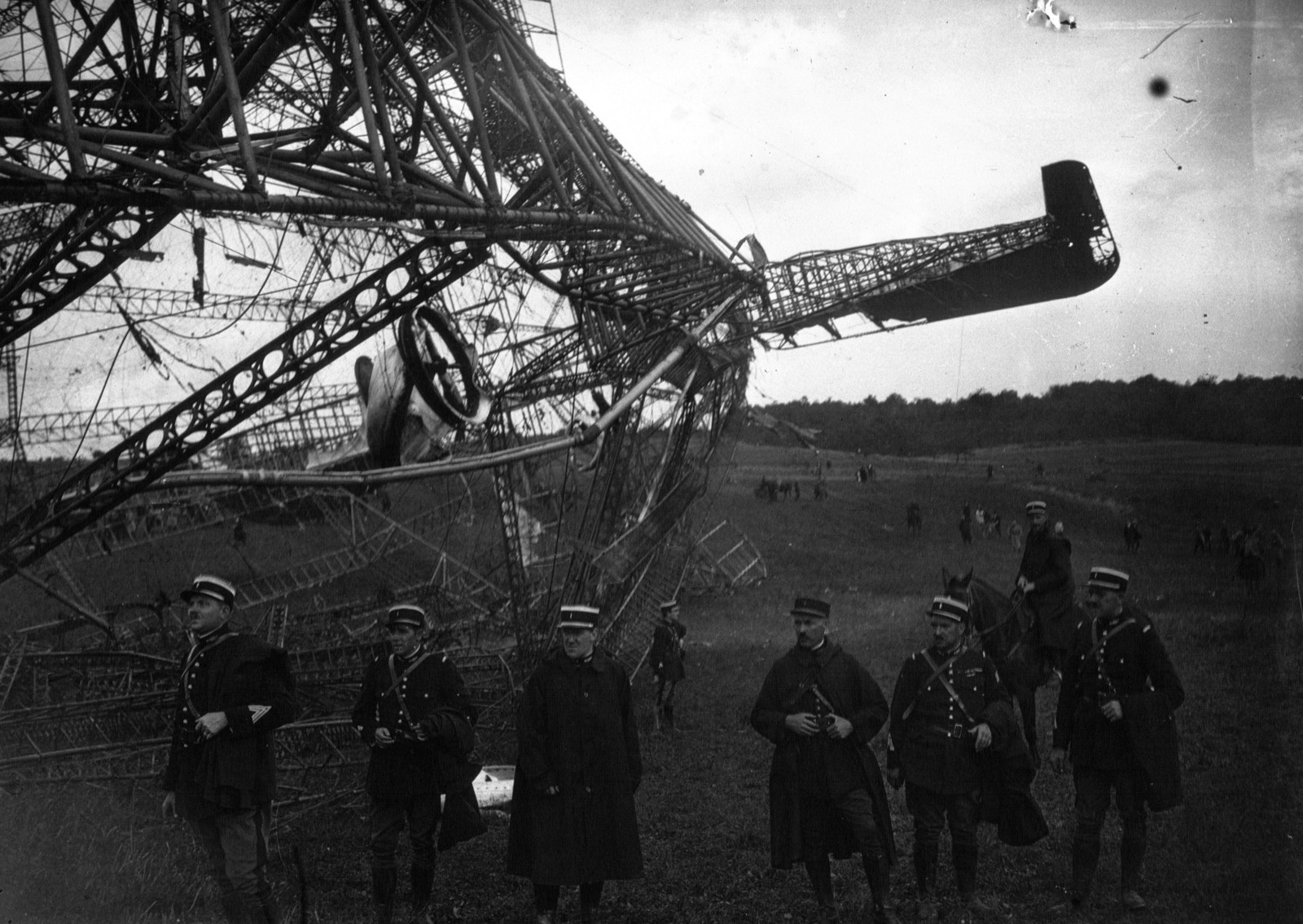 Six French gendarmes in képis stand in a field in front of the crushed girders of an airship, with a control wheel still hanging in the wreckage behind them