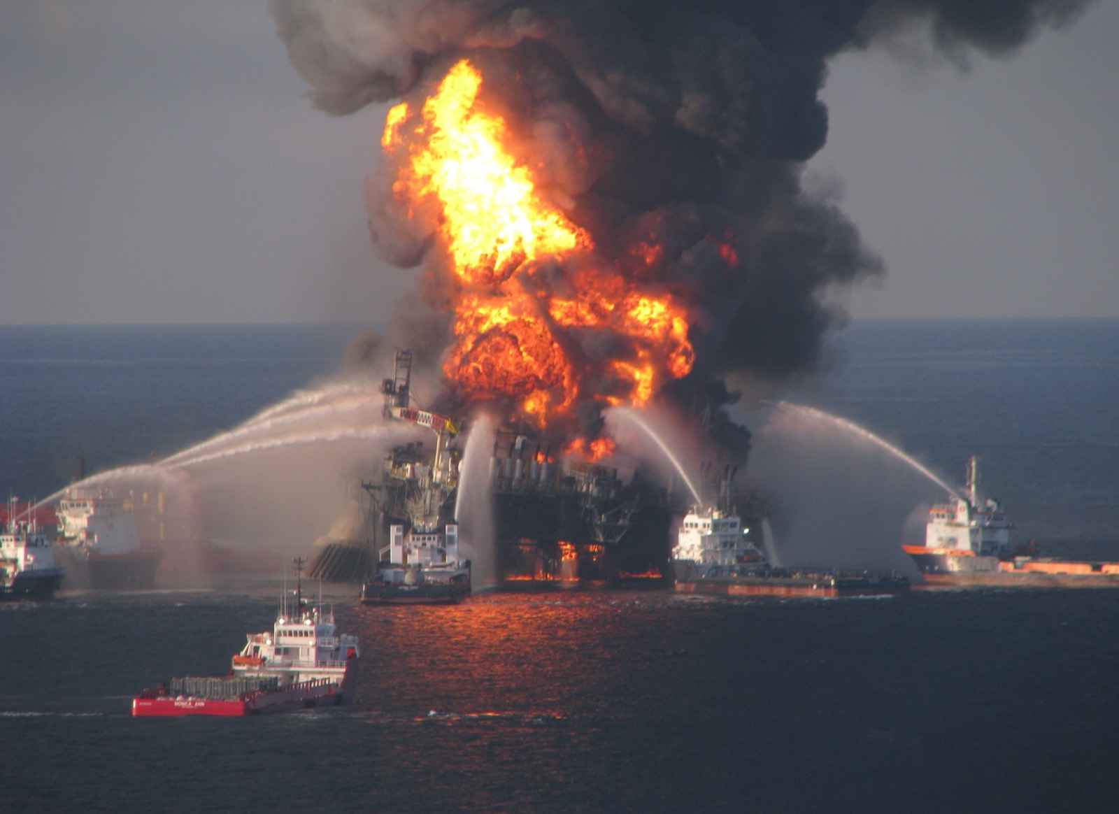 The Deepwater Horizon burning at sea, flame and black smoke rising from the derrick, with fire boats on either side playing water onto it