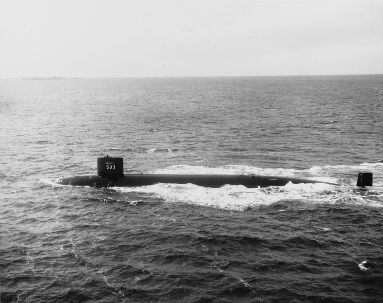 A long black submarine with the number 593 on its sail runs on the surface, water breaking white along its hull, under a pale sky