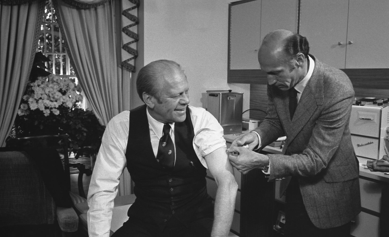 Gerald Ford in shirtsleeves and a waistcoat sits with his left sleeve rolled up, smiling, as a man in a tweed jacket gives him an injection in a small office with flowers in the window