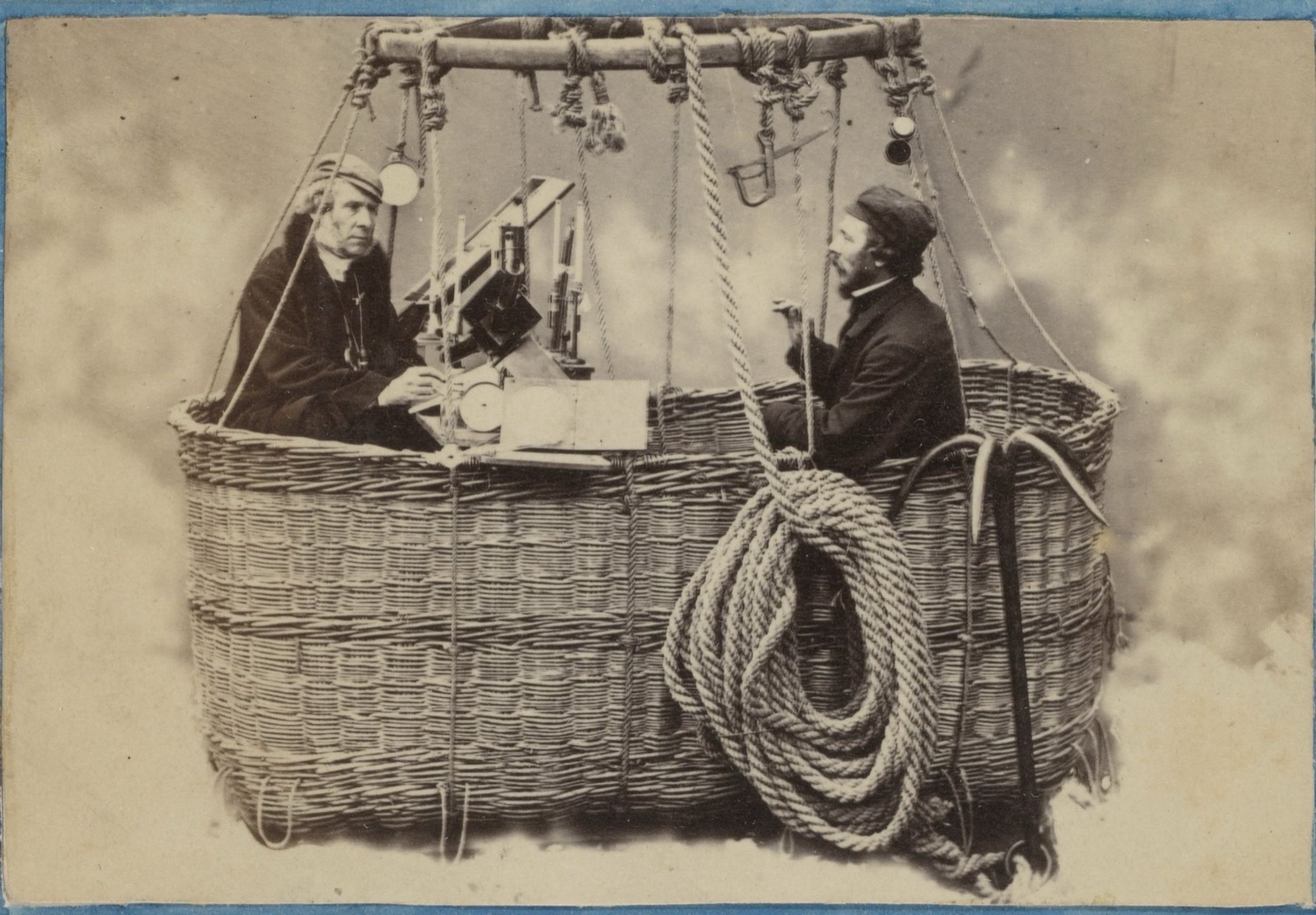A sepia photograph of two men in a wicker balloon car hung from a wooden hoop: an older man in a cap at a board of instruments, a bearded man in a cap at the other end by a coiled rope and a grapnel, against a painted sky