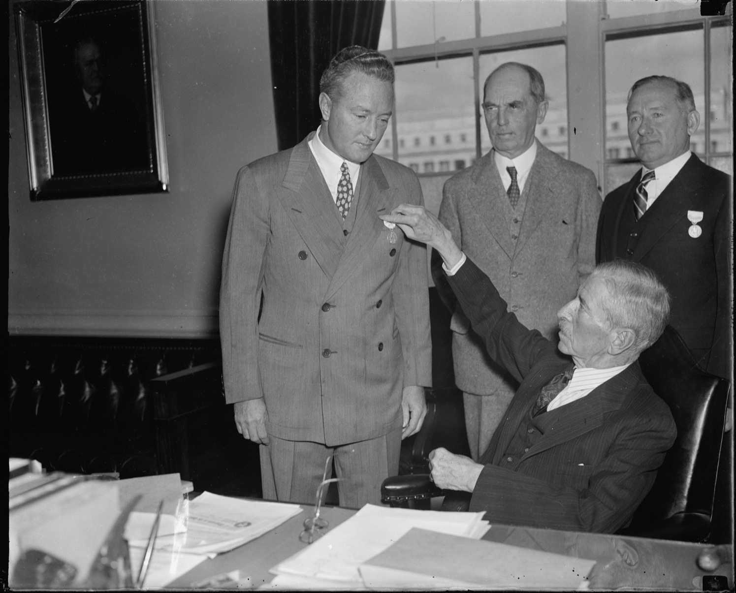 An elderly seated official pins a medal to the lapel of a man in a double-breasted suit while two other men stand behind, one of them wearing a medal of his own, in an office with tall windows