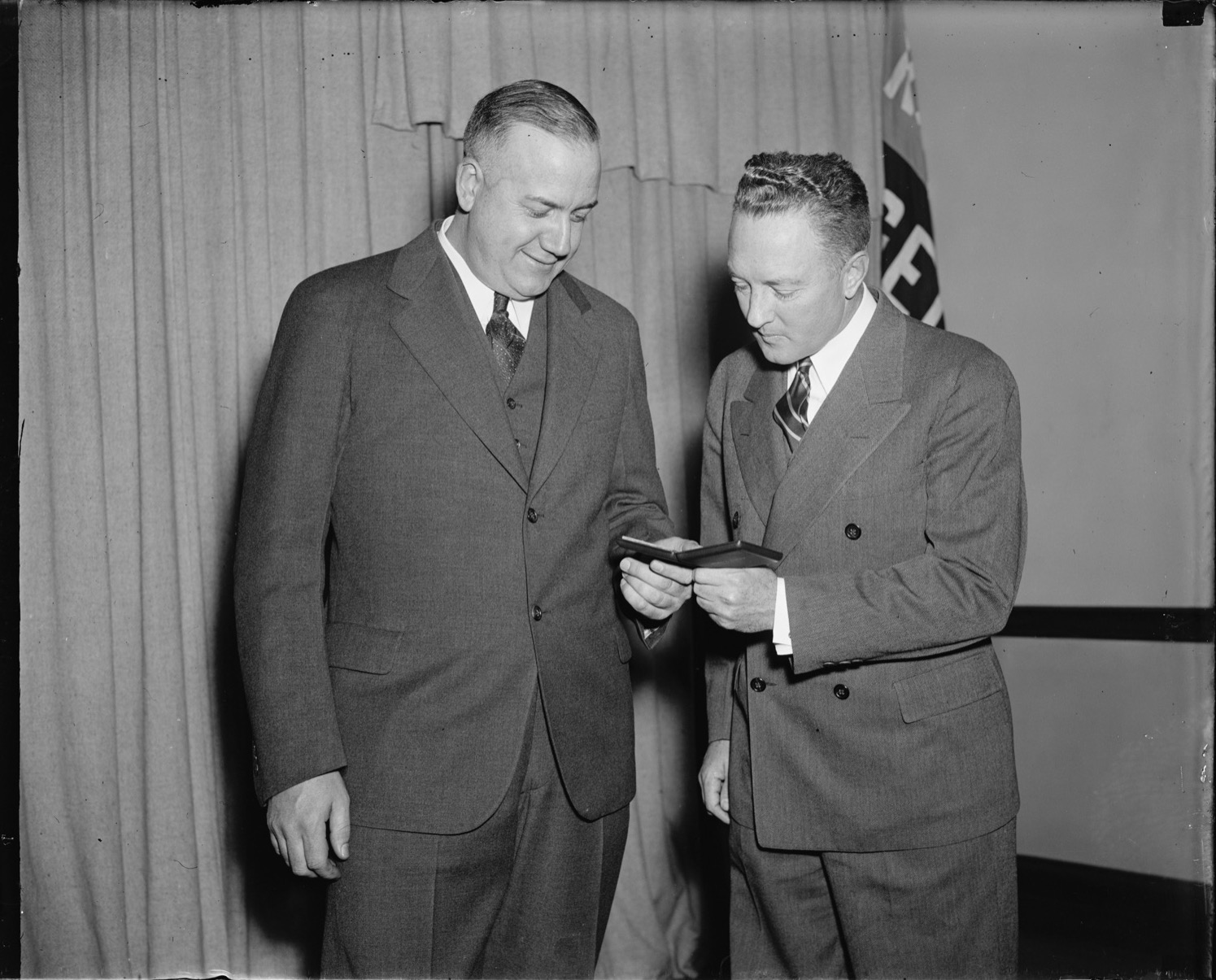 Two men in suits standing before a curtain, the larger man on the left smiling down at a small open case that the slighter man on the right holds and examines