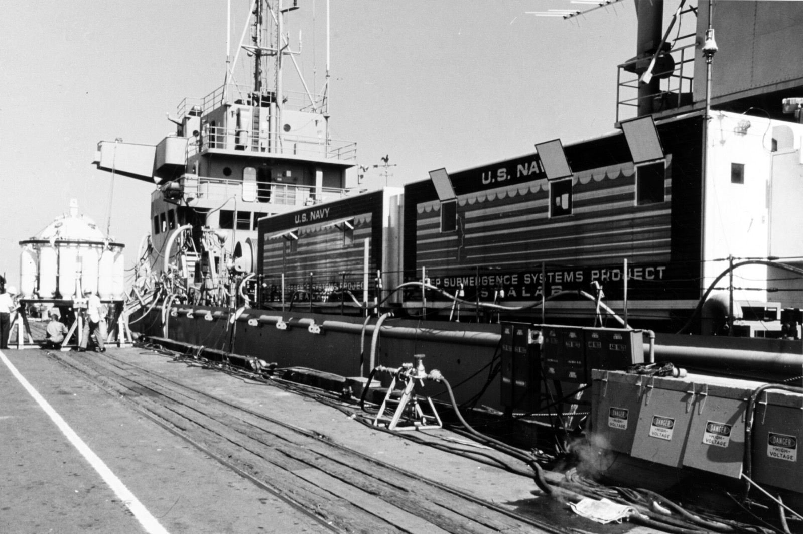 A grey naval ship alongside a pier with two long striped vans on its deck marked Deep Submergence Systems Project, and a spherical capsule standing on the pier at left