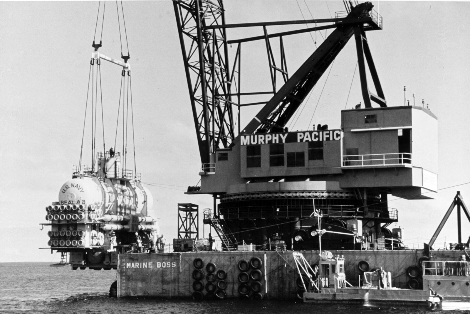 A long yellow cylinder marked US Navy Sealab, hung from the cables of a huge floating crane marked Murphy Pacific and Marine Boss, is lowered into calm water