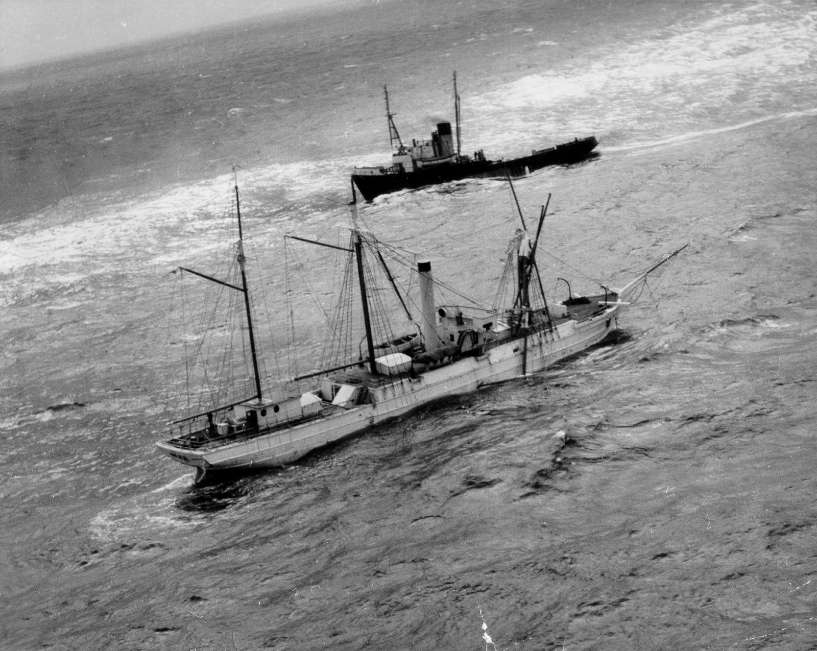 Aerial photograph of the Bear low in a rough sea with her foremast collapsed and a tug standing off