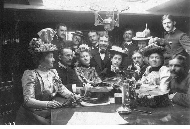 Officers and guests around a table under a hanging lamp in the Bear's wardroom, 1899