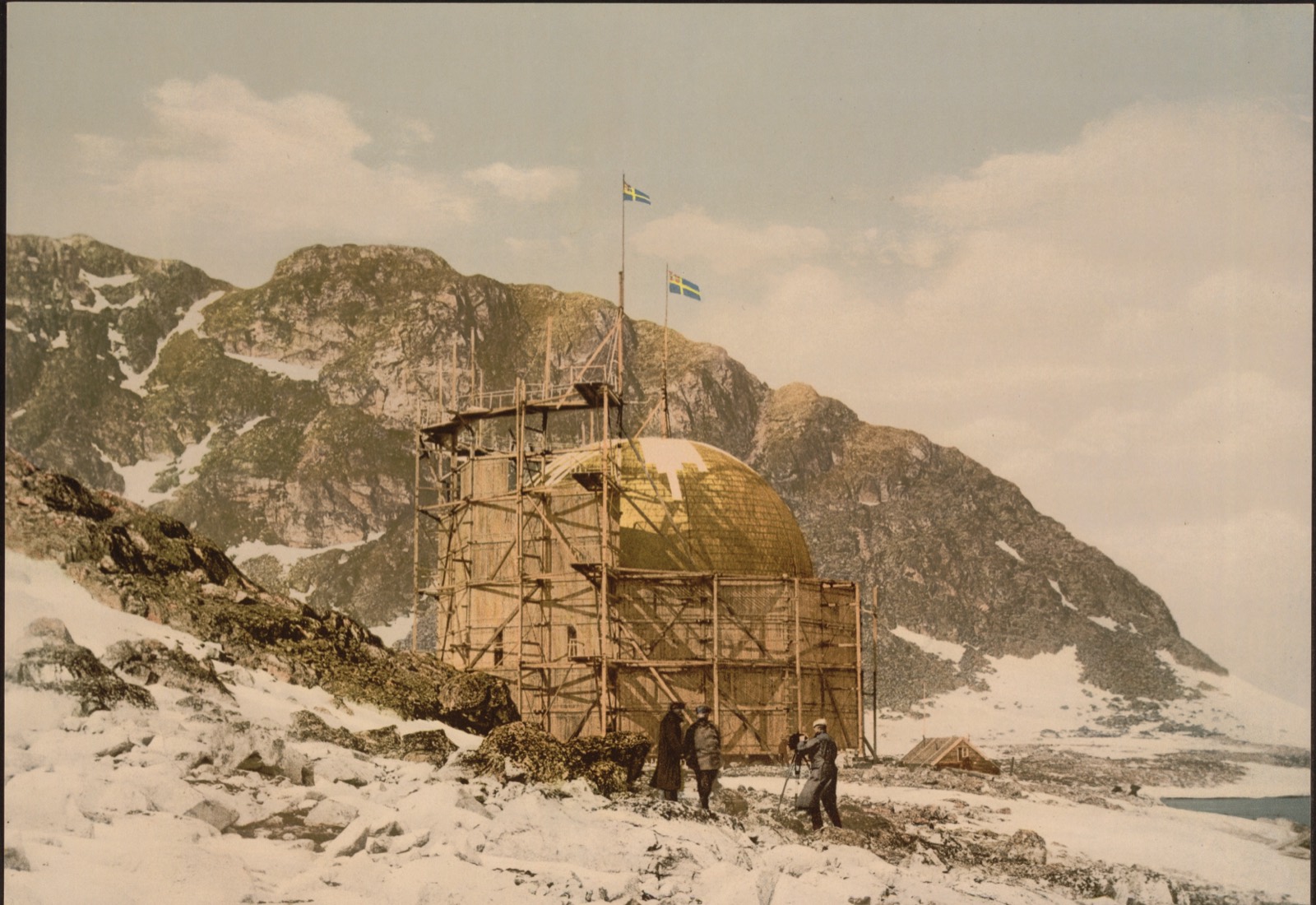 Colour print of a tall timber scaffold on a stony shore, the yellow dome of a balloon showing above its walls, two Swedish flags on poles, mountains behind