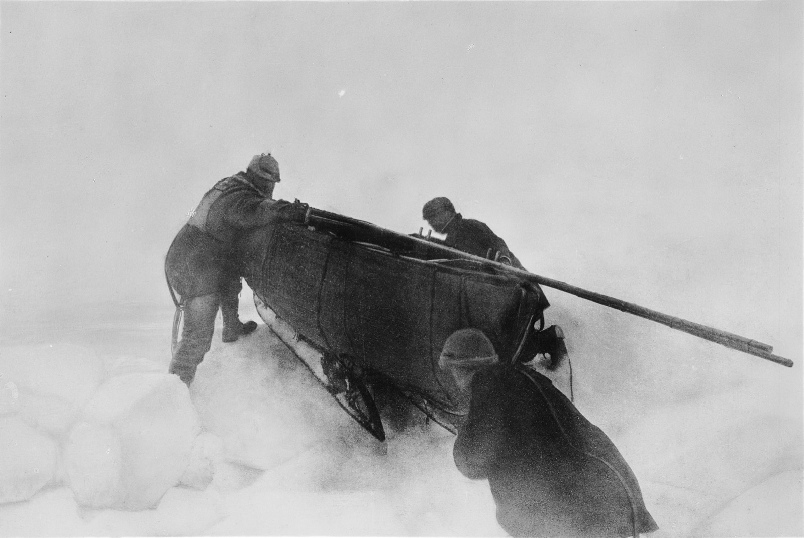 Three men in a white haze heave a loaded sledge with the boat lashed on it over blocks of pressure ice, one pulling from the front, two pushing behind