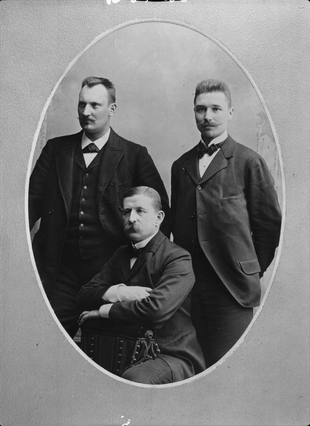 Studio portrait in an oval mount: two young men standing in dark suits, an older man with a heavy moustache seated between them with arms folded