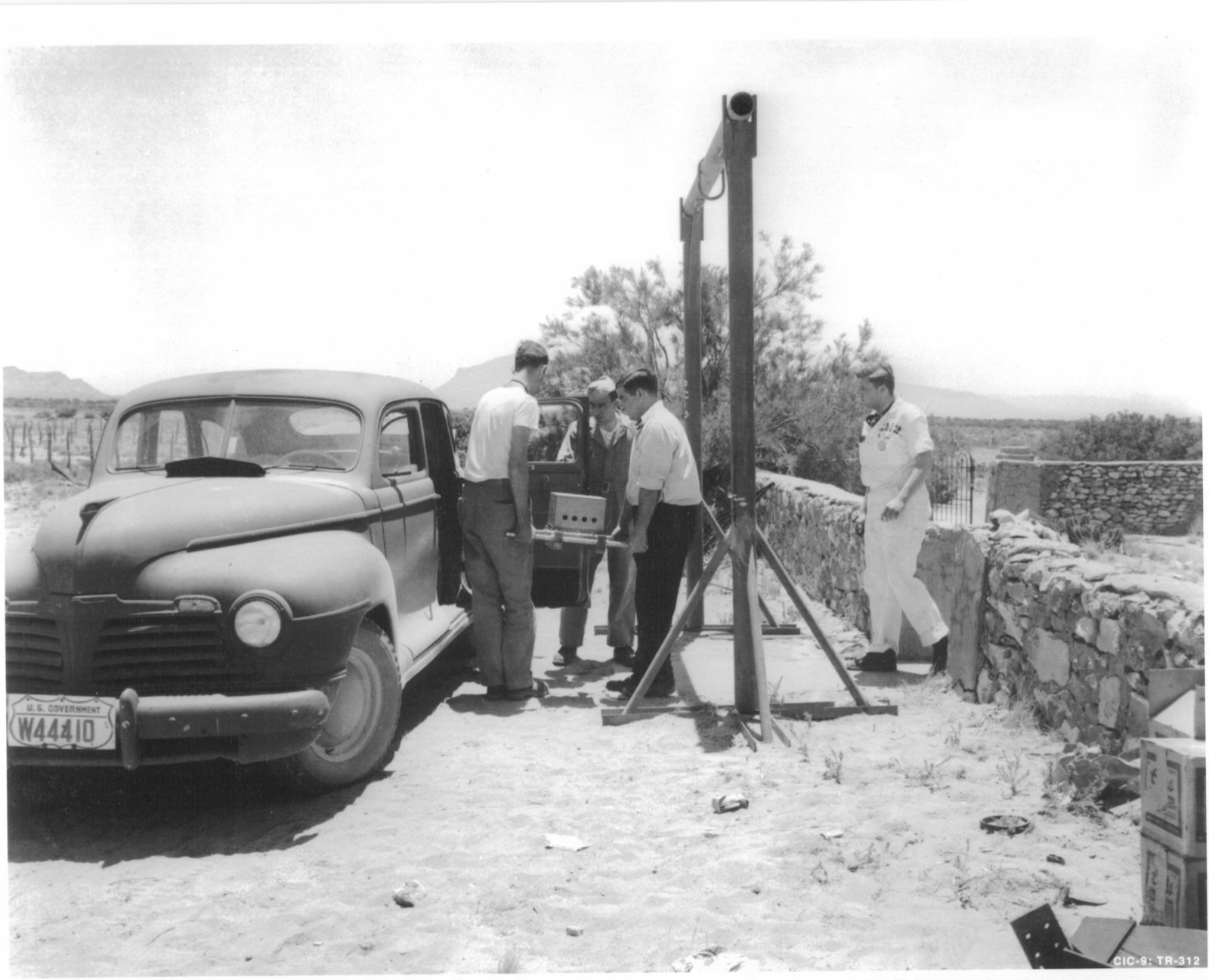 Men lifting a small boxed assembly into the open rear door of a 1940s sedan with a US Government number plate, beside a steel hoist frame and a low stone wall in the desert