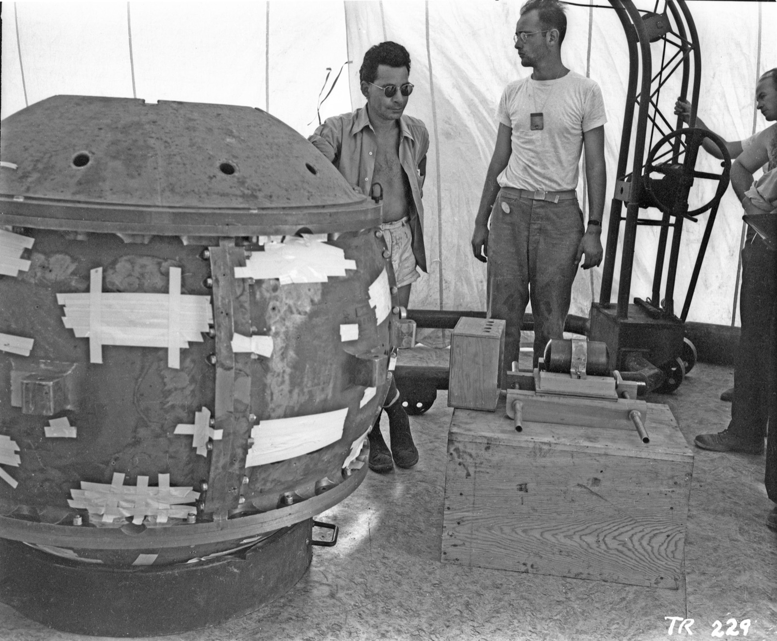 Two men beside a large riveted sphere studded with tape and cables inside a canvas tent; the nearer man wears sunglasses and an open shirt, the other a white T-shirt with a badge