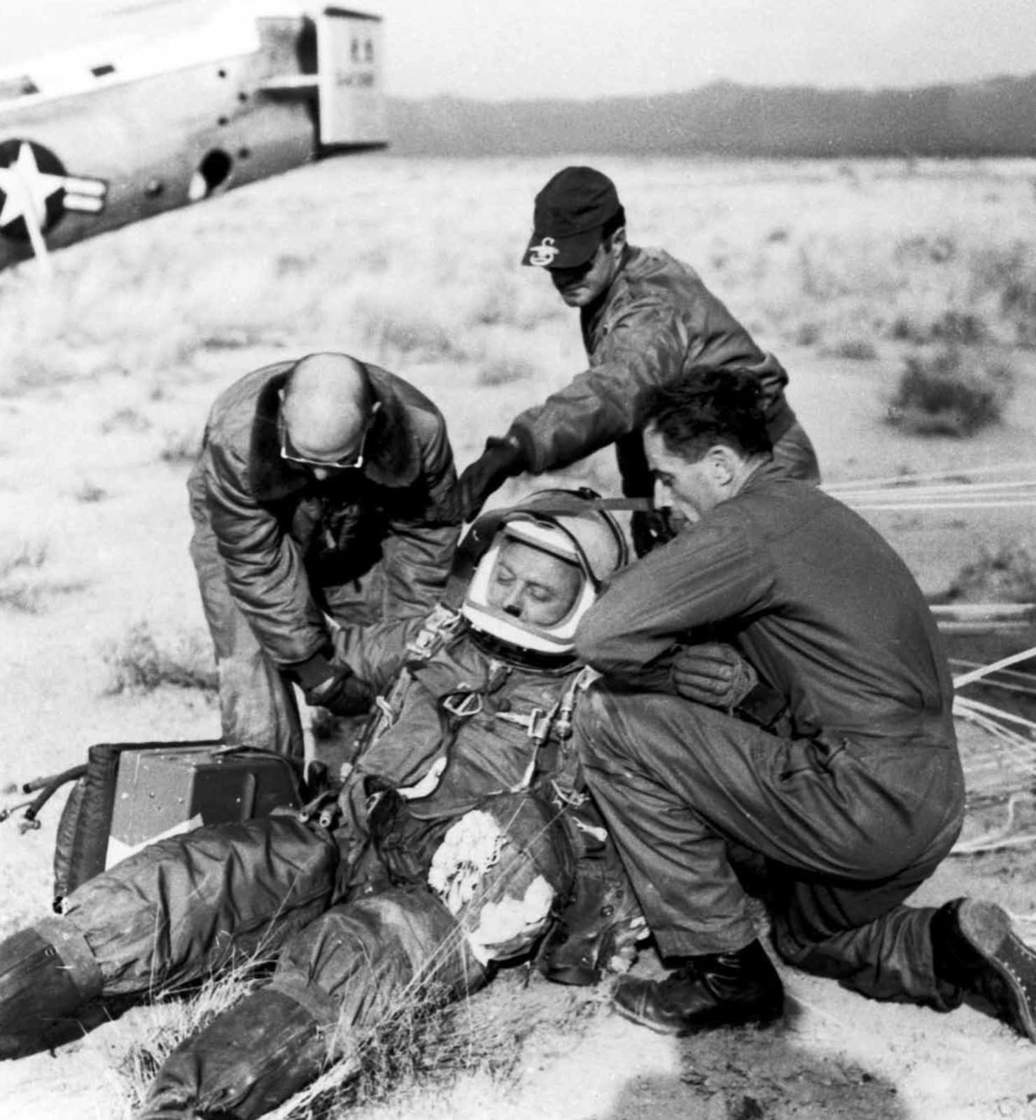 A man in a pressure suit sits on the desert ground, his helmet off and his face drawn, while three crewmen in fatigues crouch round him unbuckling his harness and equipment