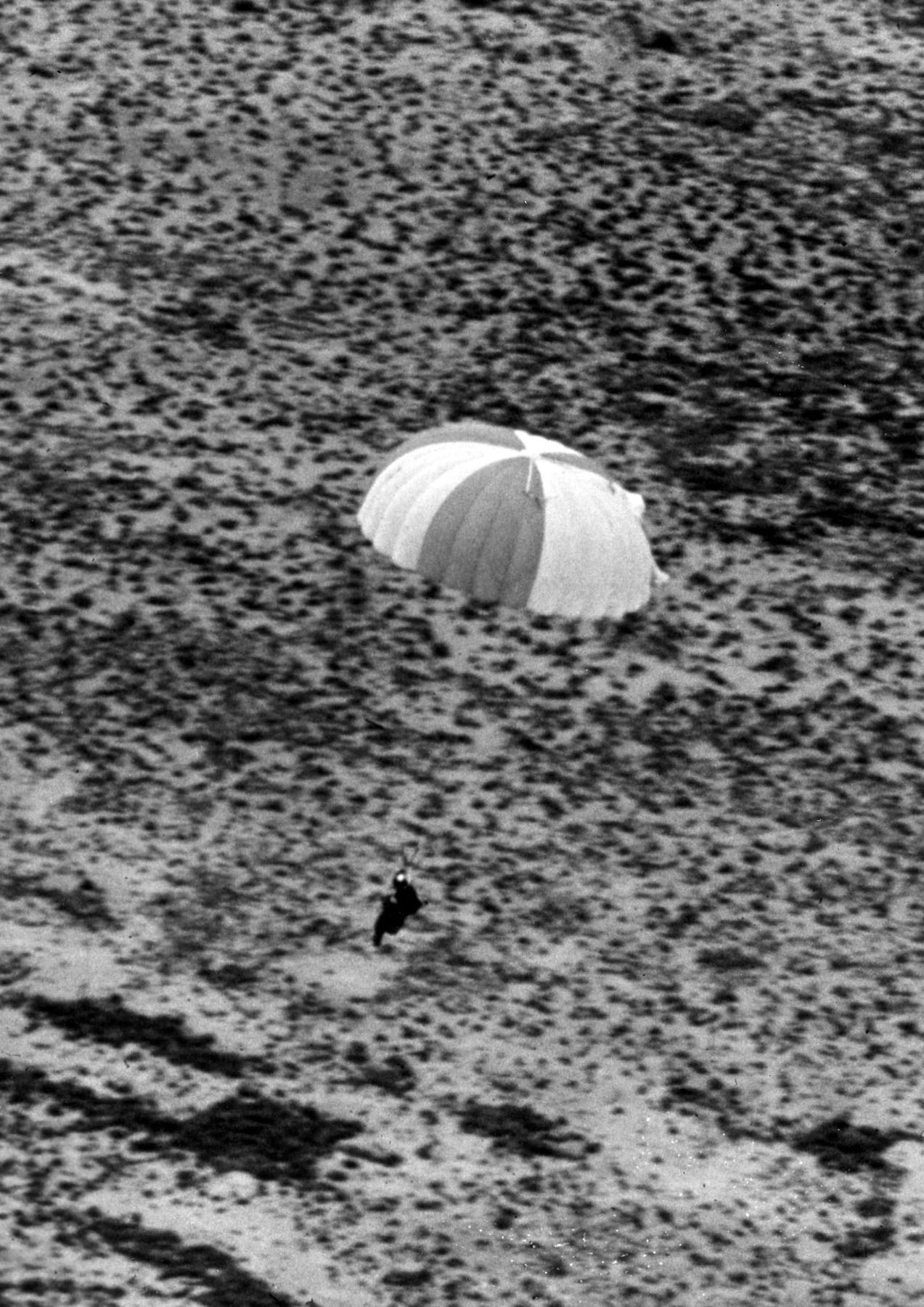 A parachutist in a bulky suit and helmet coming down under a round canopy a few feet above flat scrub desert, his legs bent for the landing