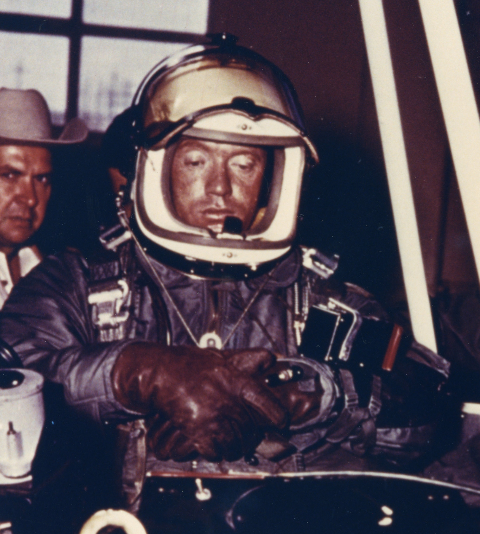 A man in a laced partial-pressure suit, a sealed helmet and a bulky parachute harness, instrument boxes strapped to his chest, looking down at the equipment