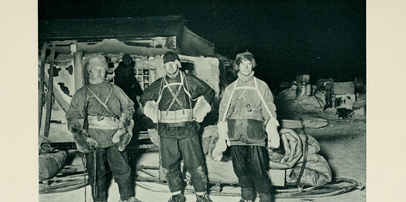 Three men in canvas wind-clothes and harness standing in front of a snow-covered hut in the dark, with laden sledges at their feet, lit by flash