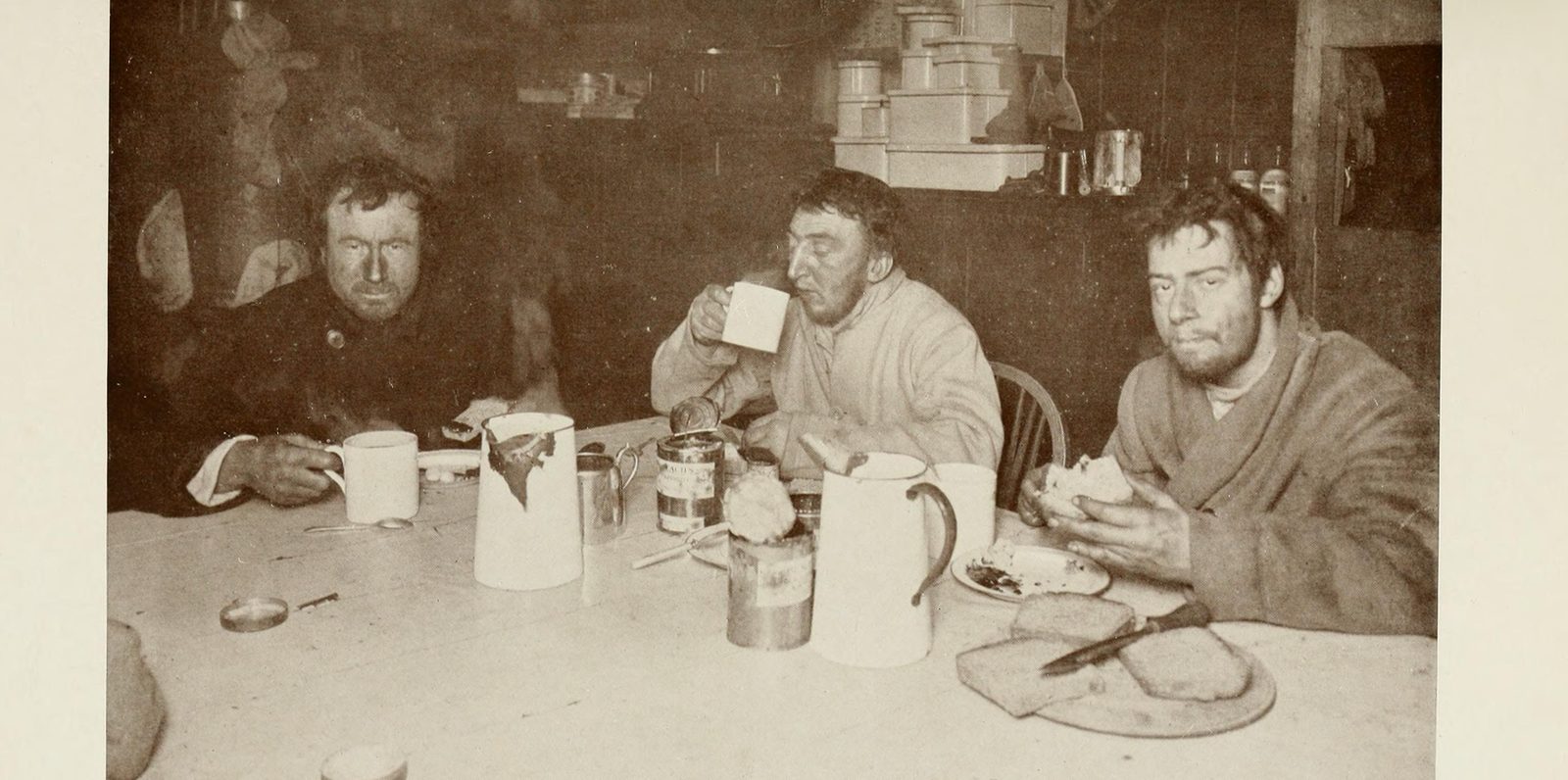 Three gaunt men with wind-burnt faces sit at a table in a wooden hut eating and drinking from mugs, with jugs, tins and a loaf in front of them