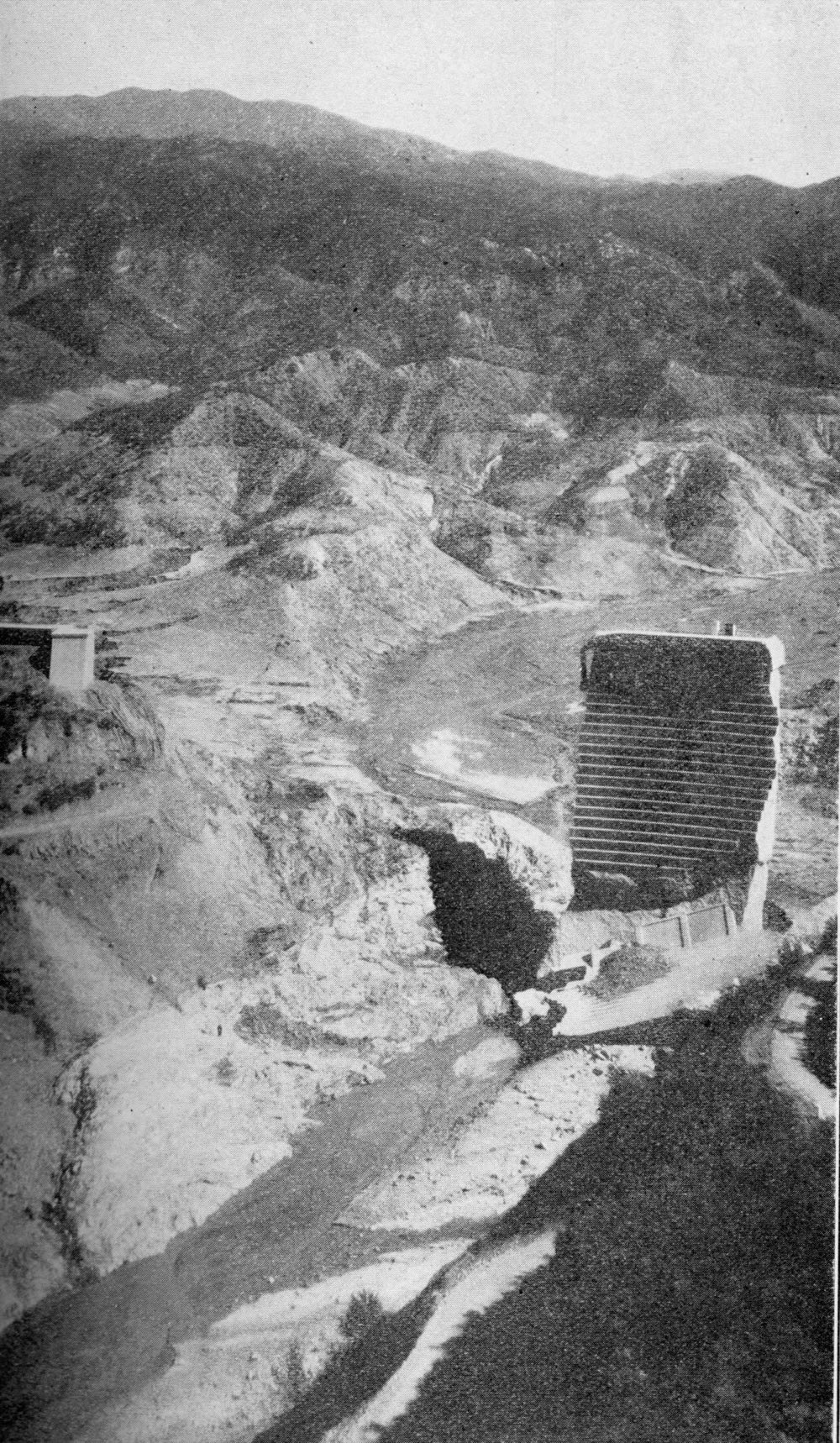 A view down into a scoured canyon with a single tall stepped block of concrete standing on the right, bare eroded hillsides on both sides, a broken wing wall on the left skyline and a stream running through the wreckage