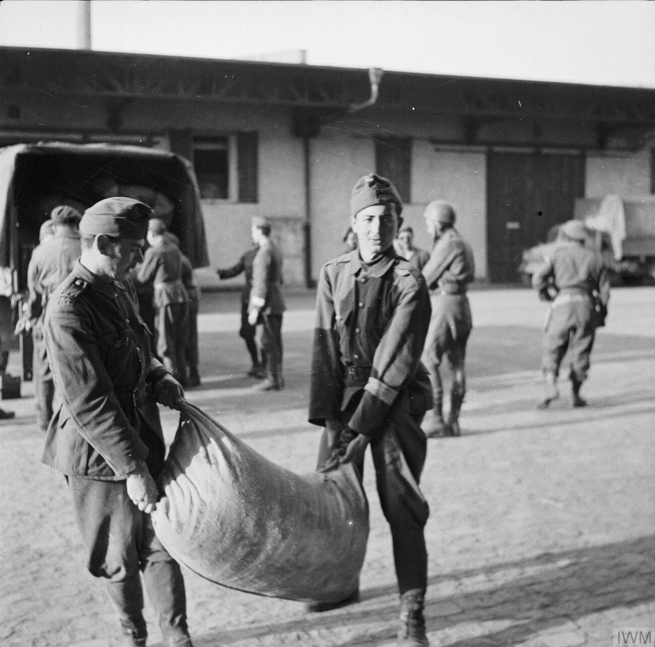 Two German soldiers carry a heavy sack between them across a yard, with other soldiers and a lorry behind them