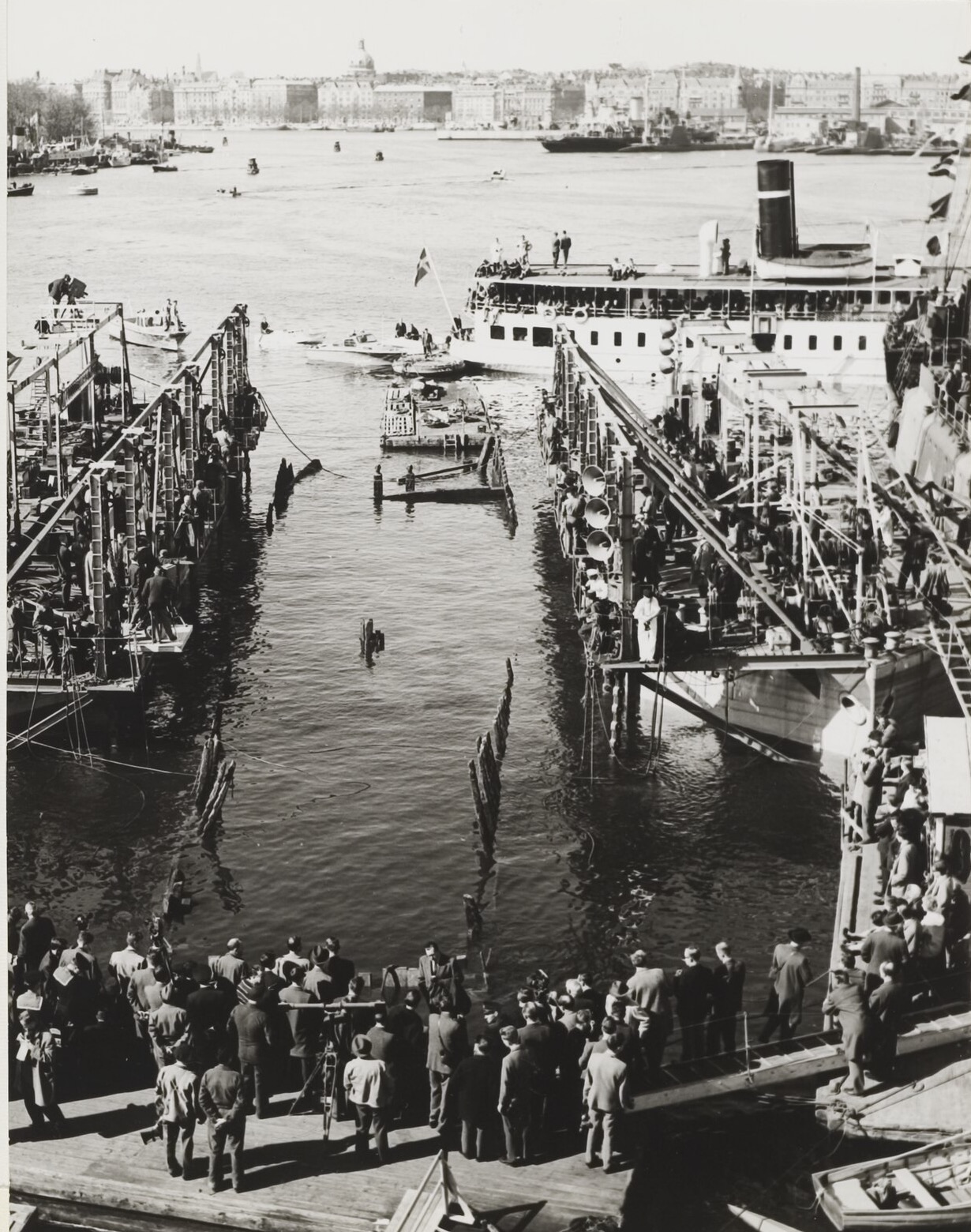 Two lifting pontoons with gantries on either side of a stretch of harbour water, the stumps of the Vasa's timbers just breaking the surface between them, a crowd watching from a quay in the foreground and a passenger steamer beyond