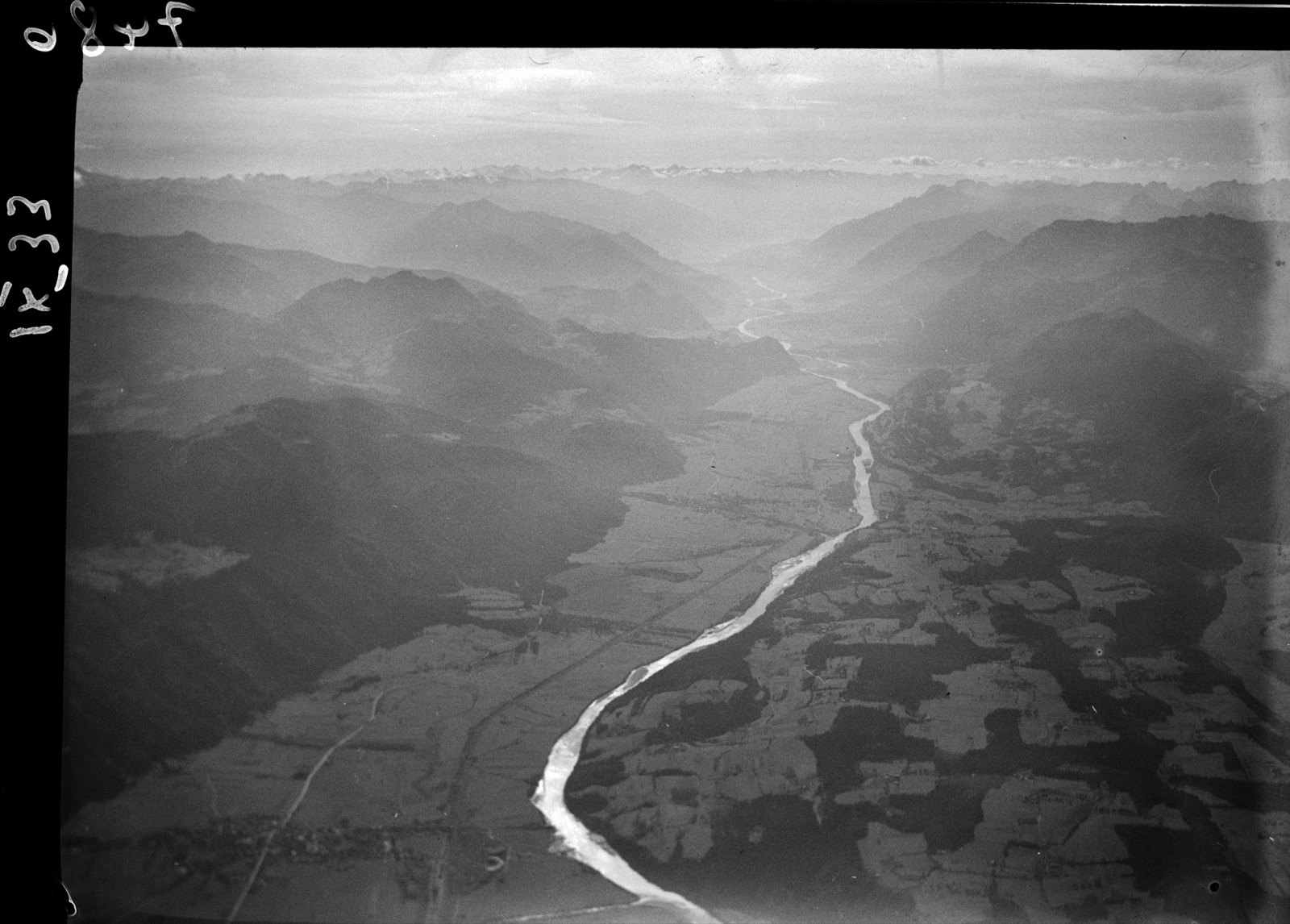 Aerial photograph of the Inn valley in the Tyrol, the river winding between dark mountains with a town at the lower left and snow peaks on the horizon