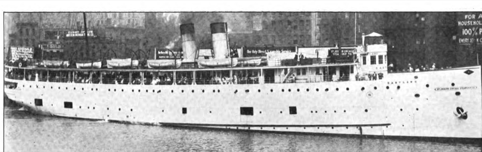 The Eastland lying at a Chicago wharf a few weeks before the accident, seen broadside from the river: a long white hull with a row of round ports close above the water, open gangway doors in the side, lifeboats along the upper deck and warehouses behind