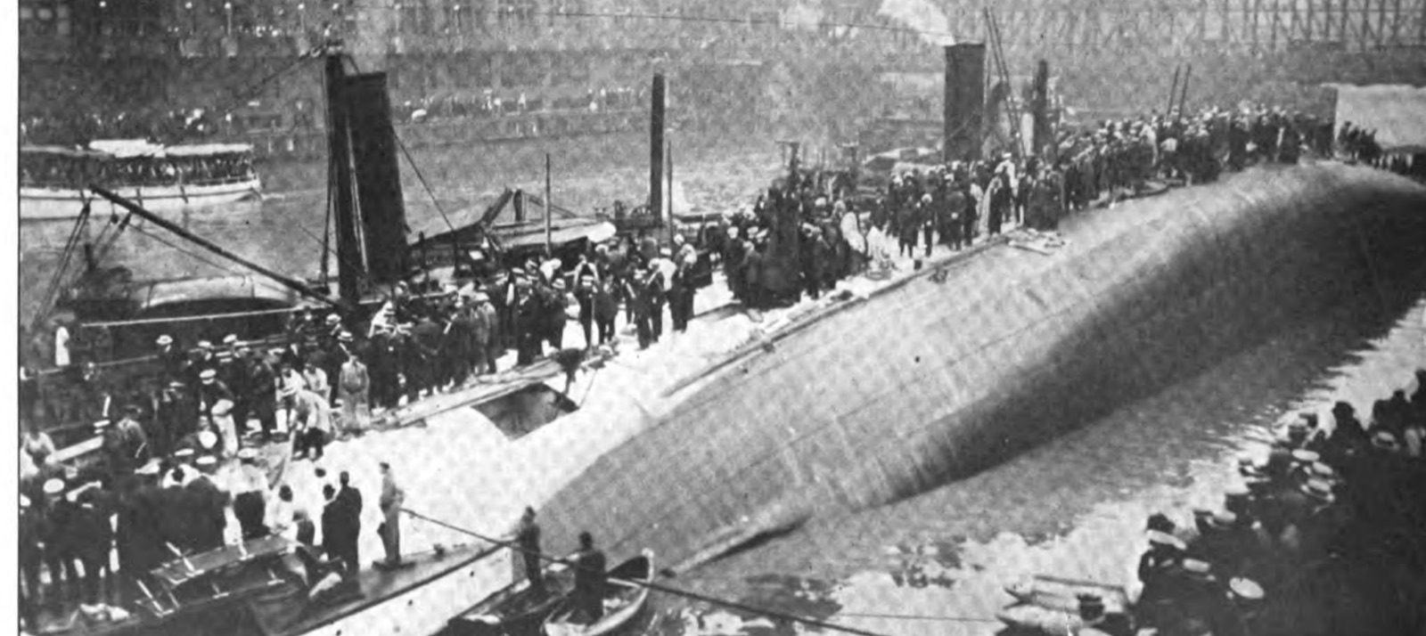 The Eastland on her side in the Chicago River immediately after capsizing, her starboard hull rising out of the water like a white whale's back with rescuers standing on it, crowds along the wharf and a bridge behind, small boats in the water