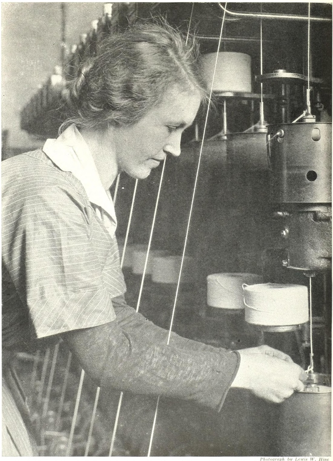 A woman in a striped work dress, seen in profile, guiding fine wire from spools through an insulating machine at the Hawthorne Works